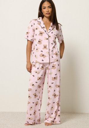 Femme portant un ensemble de pyjama rose clair avec des imprimés d'ours en peluche, haut court boutonné à manches courtes et pantalon à jambes larges, debout pieds nus sur un sol moquetté.