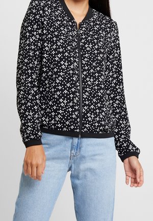 Femme portant une veste noire zippée avec des motifs floraux blancs et un jean bleu clair, montrée du cou aux hanches sur un fond uni.