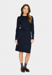 Robe longue navy à manches longues, avec un col rond et des poignets côtelés, présentant une taille cintrée et une jupe droite. Associée à des chaussettes tricotées grises et des chaussures marron.