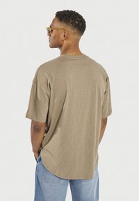 Olive green oversized t-shirt with a round neck, soft texture, and a curved hem. Worn with light blue jeans and sunglasses.
