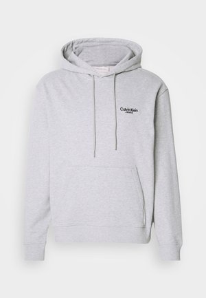 Gaiši pelēks džemperis ar kapuci no mīksta auduma, ar kenguru kabatu, aukliņām un melnu izšūtu logotipu uz krūtīm.