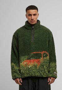 Grüner Fleece-Pullover mit einem Viertel-Reißverschluss, der eine strukturierte Oberfläche und eine Grafik eines orangefarbenen Autos vor grüner Kulisse auf der unteren Vorderseite aufweist.