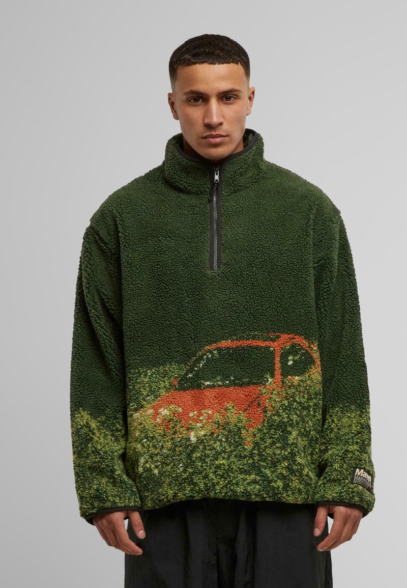 Grüner Fleece-Pullover mit einem Viertel-Reißverschluss, der eine strukturierte Oberfläche und eine Grafik eines orangefarbenen Autos vor grüner Kulisse auf der unteren Vorderseite aufweist.