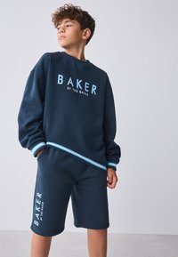 Marineblå strikket sweatshirt med "BAKER"-logo i blå, parret med matchende marineblå shorts. Begge har kontrasterende blå detaljer ved manchetterne.