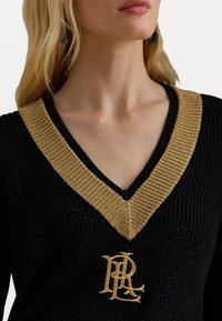 Melna adīta džemperis ar V veida kakla izgriezumu un zeltītām ribām pie kakla, ar izšutu zelta emblēmu centrā.