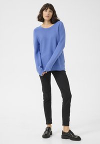 Blauer strukturierter Pullover mit langen Ärmeln, kombiniert mit schwarzen Slim-Fit-Hosen und schwarzen Loafers. Neutraler Hintergrund.