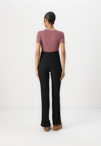 Juodos aukšto liemens kelnės su raibuliuojančia tekstūra, derinamos su trumpomis rankovėmis mauve spalvos palaidine su banguotais kraštais. Modelis stovi atsisukęs atgal.