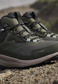 Close-up of green and black hiking shoes with textured sole and laces, resting on rocky ground outdoors.
