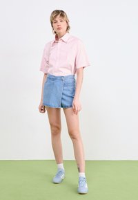 Camicia rosa chiaro a maniche corte con bottoni, abbinata a una mini gonna di jeans blu. Il modello indossa sneakers azzurri e calze bianche a mezzaluna.