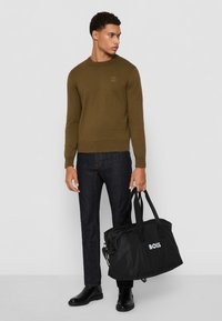 Maglione verde oliva, jeans in denim scuri e stivali di pelle nera. Il modello tiene una borsa da viaggio nera con la scritta bianca "BOSS".