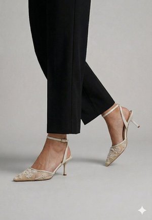 Piernas con pantalones negros recortados y zapatos de tacón altos de malla beige con bordado floral y correas en los tobillos sobre un suelo gris.