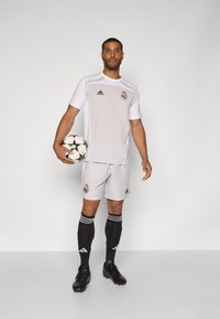 Valkoinen Adidas-jalkapallosetti mustilla yksityiskohdilla, jossa on Real Madridin logo, yhdistettynä mustiin sukkiin ja mustiin jalkapallokenkiin, pitelee kuvioitua palloa.