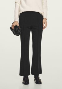 Persona con pantaloni flare neri su misura, maglione bianco, mocassini neri e una borsa intrecciata nera, davanti a uno sfondo neutro.