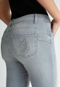 Jeans in denim grigio chiaro con design aderente, caratterizzati da cuciture decorative e strass sulla tasca posteriore. Tasche laterali visibili.