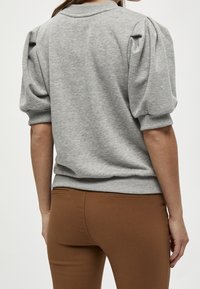 Grå tröja med puffade korta ärmar, ribbade muddar och nederkant. Buren med bruna tajta byxor som visar en slät, texturerad tygkvalitet.