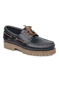 Zapato náutico de cuero negro con suela de goma marrón. Presenta ojales de metal, cordones y detalles cosidos a lo largo del empeine y la suela exterior.