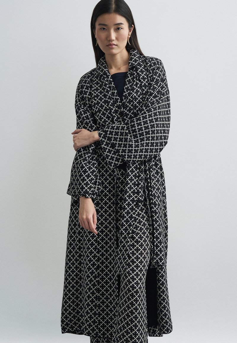 Μαύρη μακριά ρόμπα με λευκό γεωμετρικό σχέδιο, διαθέτοντας φαρδιές μανσέτες, ζωνάκι και ανάλαφρη σχεδίαση. Η υφή του υφάσματος προσθέτει βάθος.