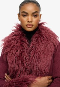 Bordeaux nepbontjas met een volumineuze kraag, gemaakt van gestructureerd materiaal, met een aansluitende zwarte coltrui eronder.