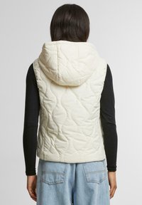 Gilet imbottito senza maniche con cappuccio, caratterizzato da un motivo trapuntato e zip frontale, abbinato a una maglia nera a maniche lunghe e jeans azzurri chiari.