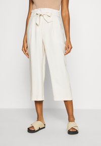 Culottes taille haute beige clair avec ceinture à nouer et plis, caractérisées par une coupe ample et une longueur raccourcie, assorties à des sandales plates beiges.
