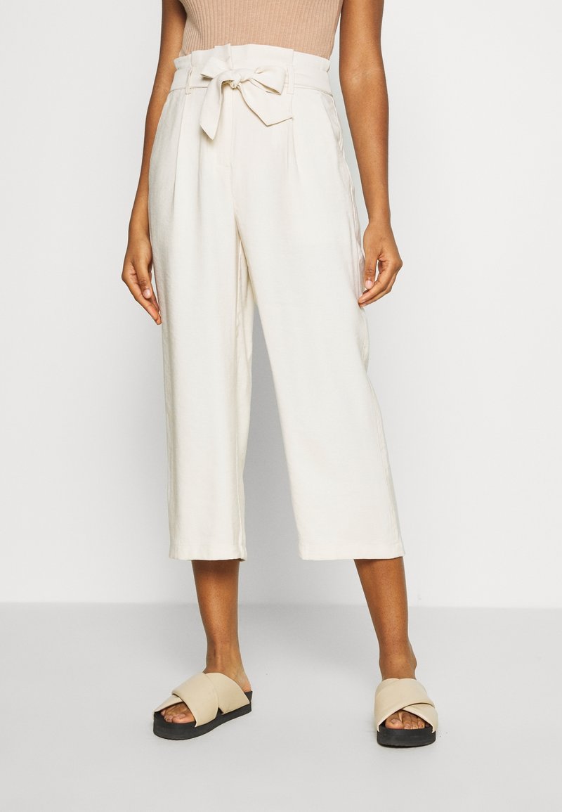 Culottes taille haute beige clair avec ceinture à nouer et plis, caractérisées par une coupe ample et une longueur raccourcie, assorties à des sandales plates beiges.
