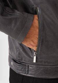 Grå läderjacka med en dragkedjeficka, slät textur, tryckknappsstängning i midjan och detaljerad sömnad.