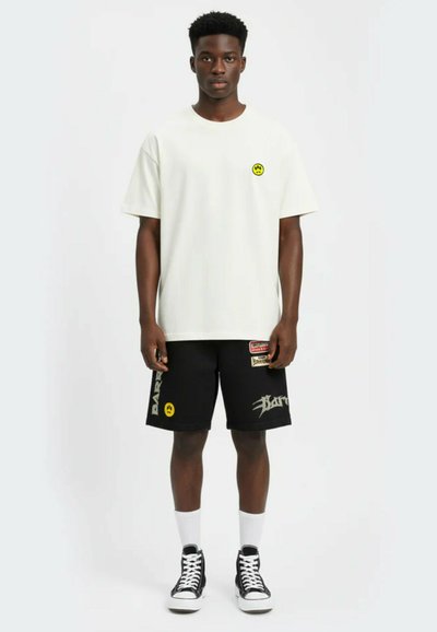 Junger Mann steht, trägt ein weißes T-Shirt mit einem kleinen gelben Smiley-Gesicht und schwarze Shorts mit mehreren Aufnähern und Logos.