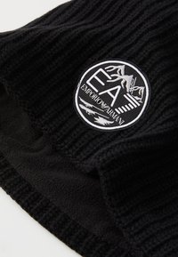 Crni rebrasti pleteni materijal s kružnom crno-bijelom oznakom Emporio Armani EA u�šivenom na njemu.