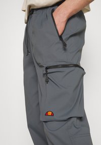 Pantalon cargo gris avec poches zippées, une poche entrouverte avec la main à l’intérieur, et un petit logo rouge et orange sur la poche inférieure.
