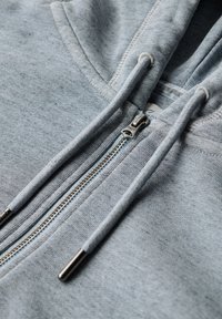 Hellgraue Zip-Hoodie aus weichem Material, ausgestattet mit einem Metallreißverschluss mit Zipper und verstellbaren Zugbändern am Kragen.