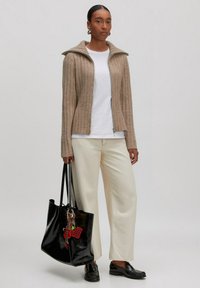 Beige gerippte Zip-Jacke, weißes T-Shirt, cremefarbene weite Hosen, schwarze Lederschuhe und eine glänzende schwarze Tasche mit bunten Anhängern.