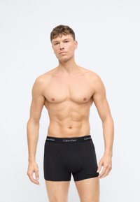 Juodos medvilnės boxer trumpikės su minkšta juostos juosta, ant kurios užrašyta "Calvin Klein" balta spalva. Modelis turi tonizuotą figūrą ir pozicionuojasi neutraliai.