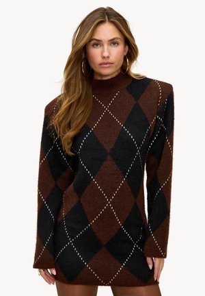 Vrouw met lang haar draagt een bruine en zwarte sweaterjurk met een ruitenpatroon en gouden creooloorbellen, kijkt naar voren tegen een neutrale achtergrond.