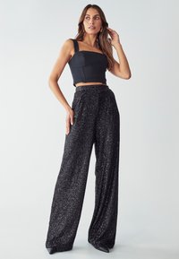 Top corto nero con spalline larghe e design strutturato, abbinato a pantaloni palazzo neri high-waisted glitterati caratterizzati da una vestibilità ampia.