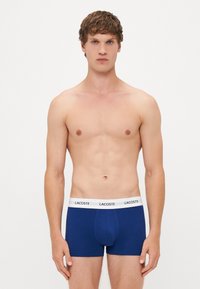Boxers en coton bleu avec une ceinture élastique blanche portant le nom de la marque "LACOSTE". Design simple, coupe ajustée et texture douce.