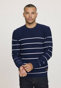 Pull en tricot bleu marine avec des rayures blanches horizontales, un col rond et des poignets côtelés. Accent en cuir près de l'ourlet. Le modèle est en position debout.
