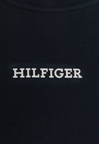 Tessuto blu scuro con una superficie liscia, caratterizzato dalla scritta "HILFIGER" in lettere bianche in rilievo e in grassetto, posizionata al centro della zona del petto.