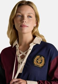 Baumwoll-Rugbyshirt mit einem Design in burgunderrot und marineblau. Verfügt über einen beigen Kragen, eine Schnürung vorne und ein gesticktes Logopatch.