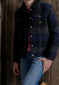 Giacca a scacchi nera e blu con colletto foderato in pelliccia, bottoni metallici, abbinata a una camicia granata e jeans blu. Sfondo in legno.