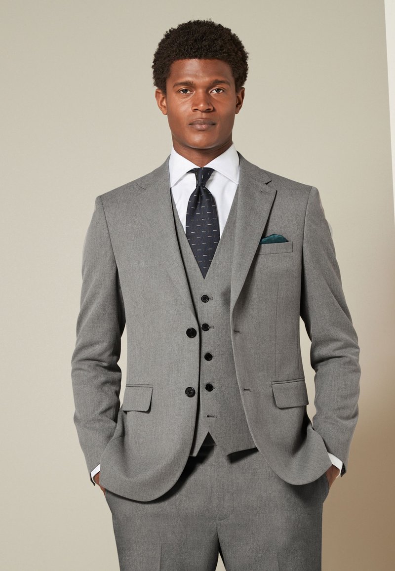 Homme portant un costume trois pièces gris clair sur mesure avec une chemise blanche, une cravate sombre à motifs et un mouchoir de poche vert, debout les mains dans les poches.