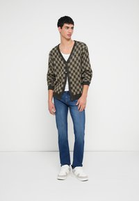 Dziany kardigan z brązowo-czarnym wzorem w kratkę, z dekoltem w serek, zapięciem na guziki i prążkowanymi mankietami; noszony na białej koszuli i niebieskich dżinsach.