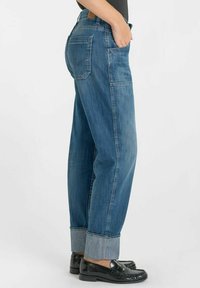 Jean en denim bleu avec une coupe décontractée, ourlets repliés et poches arrière. Porté avec des mocassins noirs. Vue de côté sur fond uni.