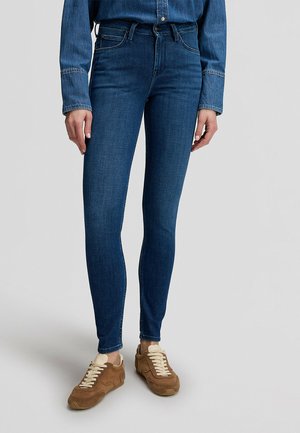 Persona che indossa jeans skinny blu scuro, camicia di jeans blu e sneakers in camoscio marrone con lacci beige, in piedi davanti a uno sfondo uniforme.