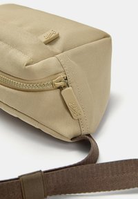 Borsa in tessuto beige con superficie testurizzata, dotata di una cerniera e di un tirante beige, attaccata a una tracolla marrone. Design robusto e compatto.