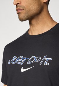 Sort bomulds t-shirt med et blåt mønstrede "JUST DO IT" logo og en hvid Nike swoosh, med rund halsudskæring og korte ærmer.