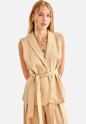 Mujer con cabello largo y rubio que lleva una blazer sin mangas beige a rayas y pantalones a juego, complementada con collares de capas coloridos y pendientes.