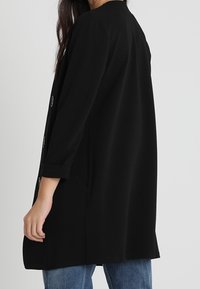 Černý dlouhý kabát s dlouhými rukávy a rovým střihem, s kulatým výstřihem, bočními kapsami a třemi knoflíky na přední straně. Jednoduchá textura.