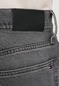 Szürke farmeranyag jellegzetes textúrával, fekete bőr címkével, amelyen a "TOMMY HILFIGER" felirat domborodik, valamint egy kis piros-fehér-kék logó díszítéssel.