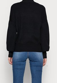 Schwarzer gestrickter Pullover mit hohem Kragen und überschnittener Schulter. Rippbündchen und Saum. Getragen über einer schmal geschnittenen blauen Jeans.