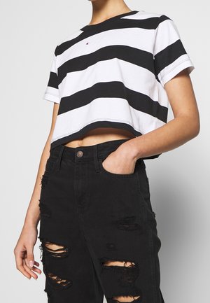 Personne portant un t-shirt blanc court à larges rayures noires et un jean noir déchiré, une main dans la poche, sur un fond uni.